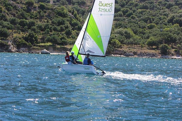 Sailor in a small sailboat with lime green and white sail on blue water near forested coastline.
