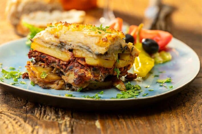Moussaka layered with eggplant, meat sauce and creamy béchamel on a blue plate, garnished with fresh herbs and served with roasted vegetables.