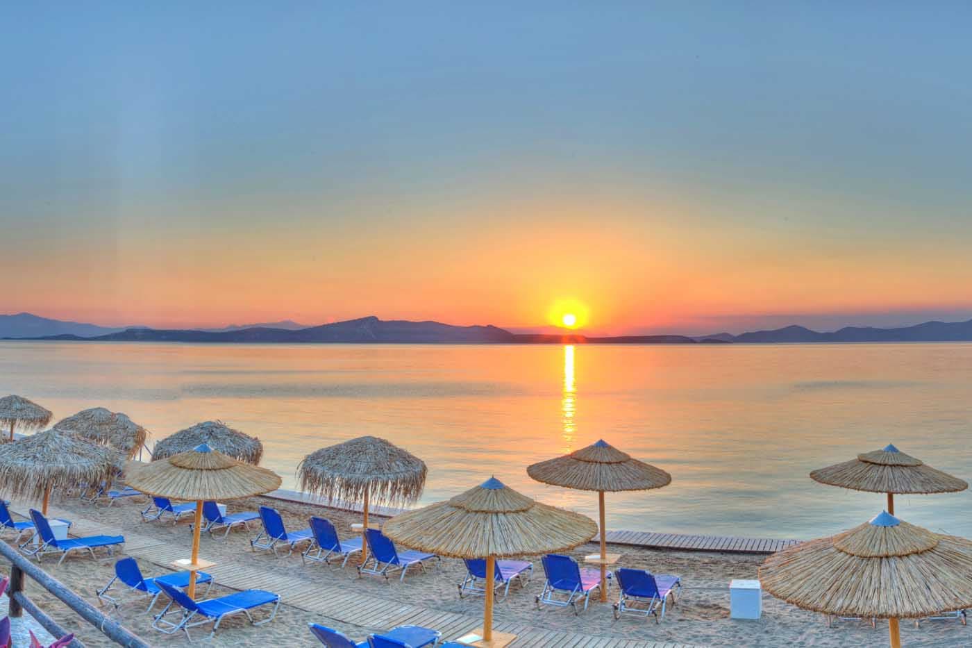Sunrise over the sea at a Greek Beach Club with blue loungers and straw umbrellas lining the sandy shore, Evia