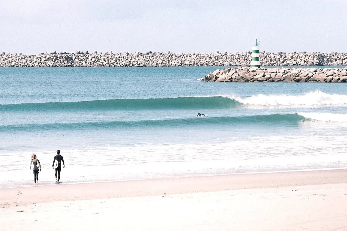 Surfers in wetsuits on sandy beach with turquoise waves and breakwater at Cabedelo, Portugal.