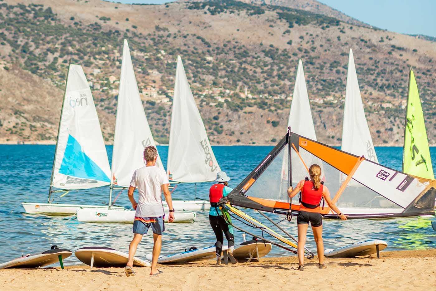 Beach Club instructors with sailing equipment and boats lined up on sandy shore, Mediterranean mountains in background